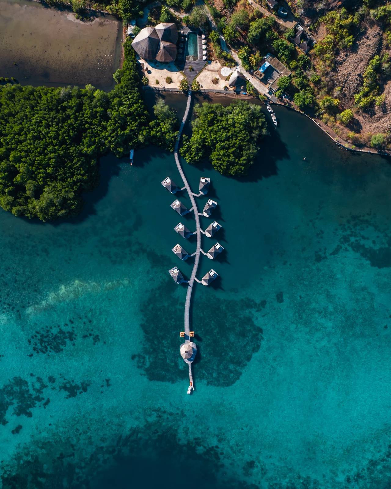 Menjaga Bay 空拍私灣：岸邊茅草頂館舍與泳池、長棧橋上成對尖頂水上小屋、末端圓亭與白船
