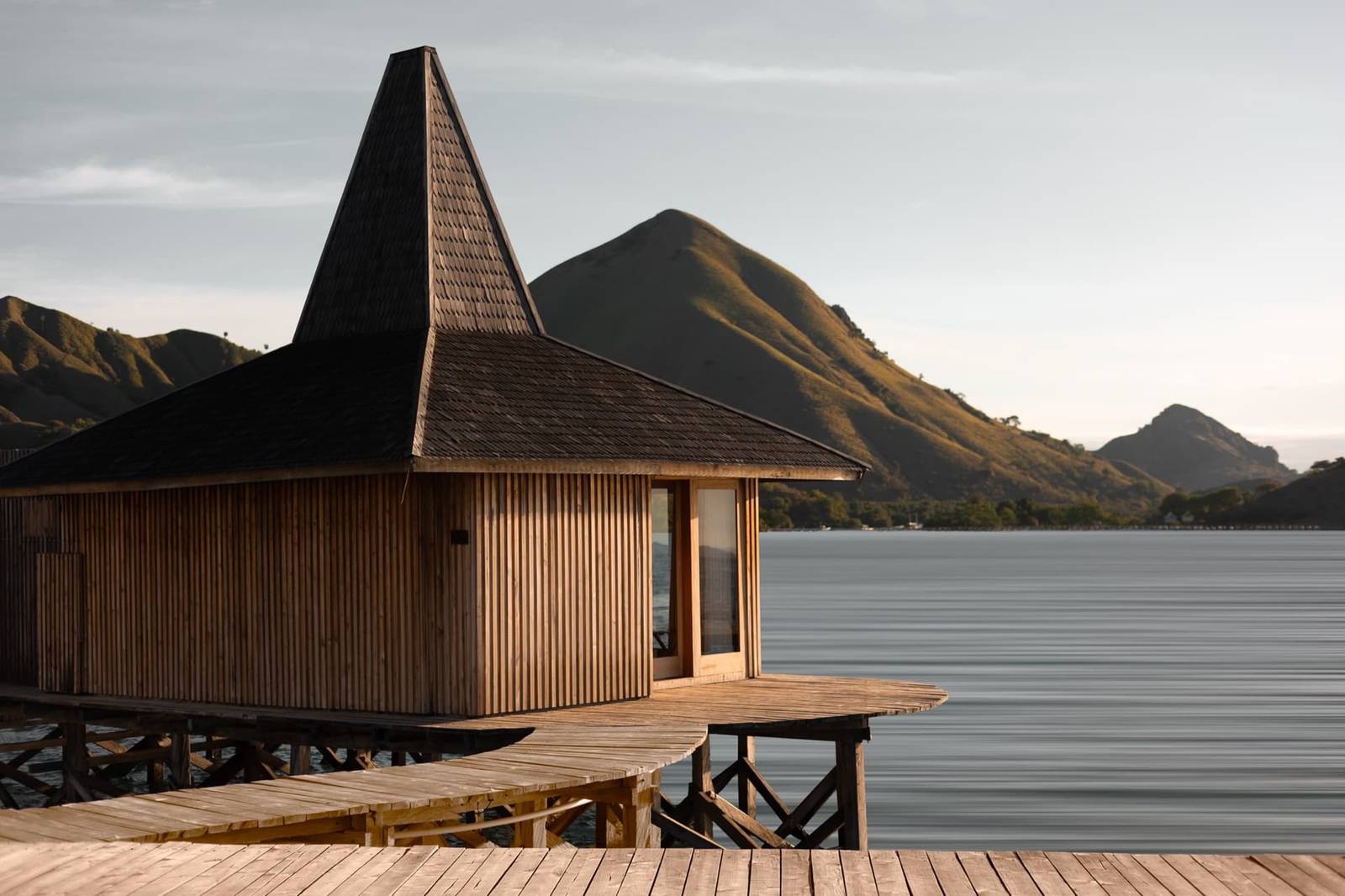 Menjaga Bay 水上木屋尖頂、豎向木條牆、棧橋通向小屋，後方乾草丘陵