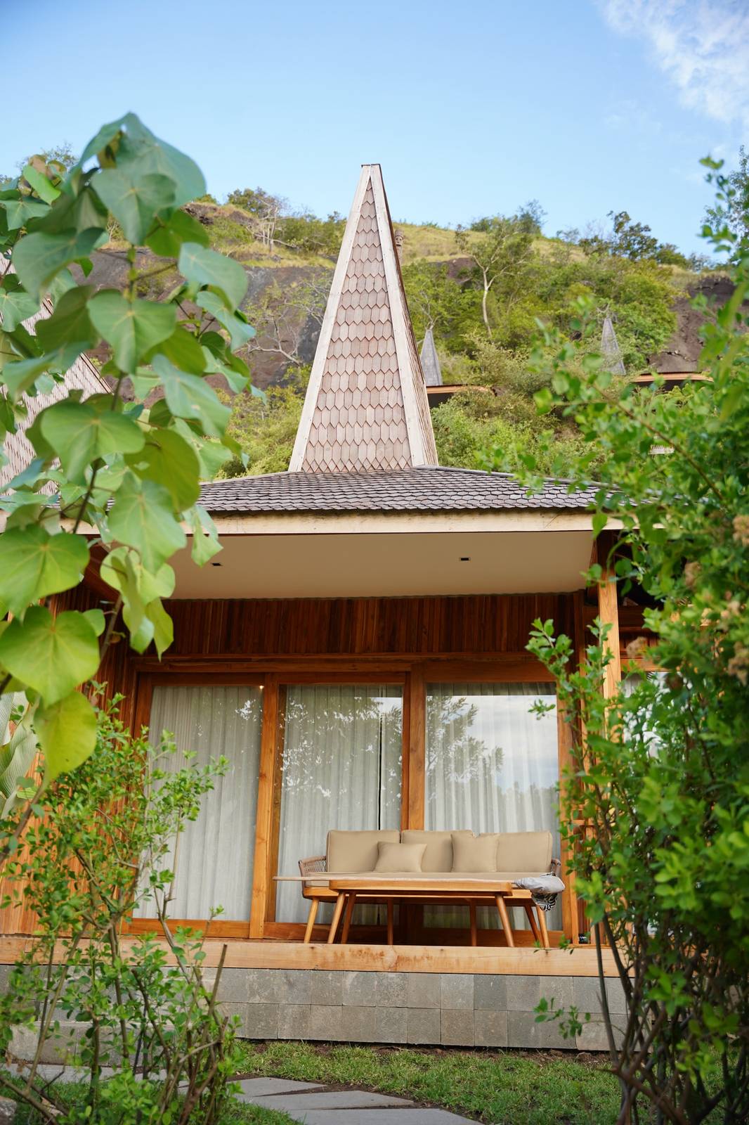 Menjaga Bay 山坡尖頂木屋、石基木陽台沙發茶几，前景熱帶葉