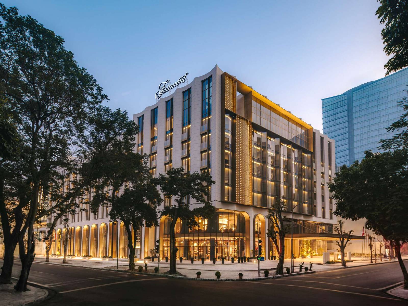 河內費爾蒙酒店黃昏街角立面,拱廊透出暖光,樓頂可見 Fairmont 草書燈箱,行道樹夾著白色量體