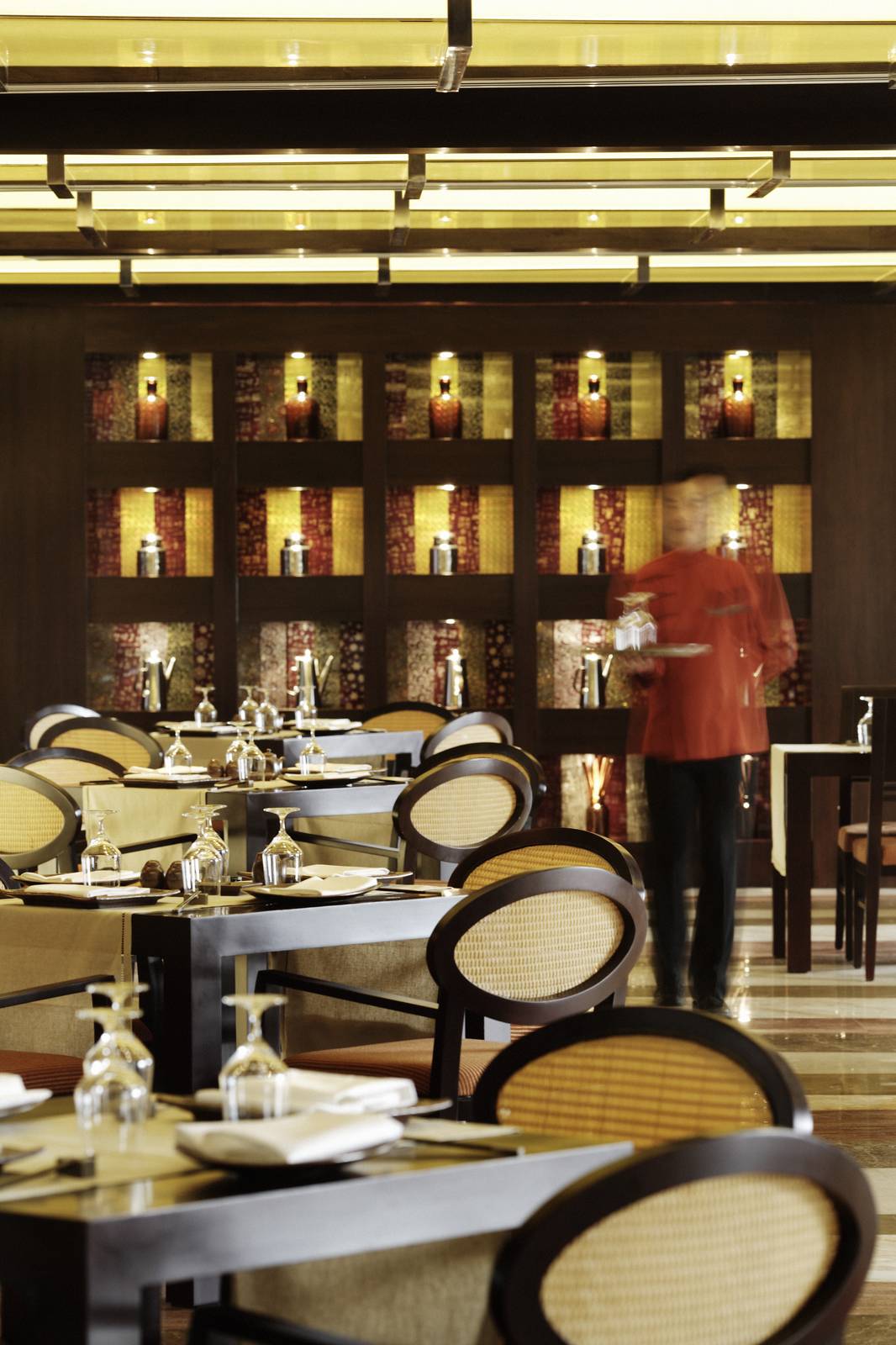 河內西湖洲際酒店 Saigon 餐廳，深木圓桌、藤編橢圓椅背與後方格狀燈龕牆，工作人員穿紅襖端托盤