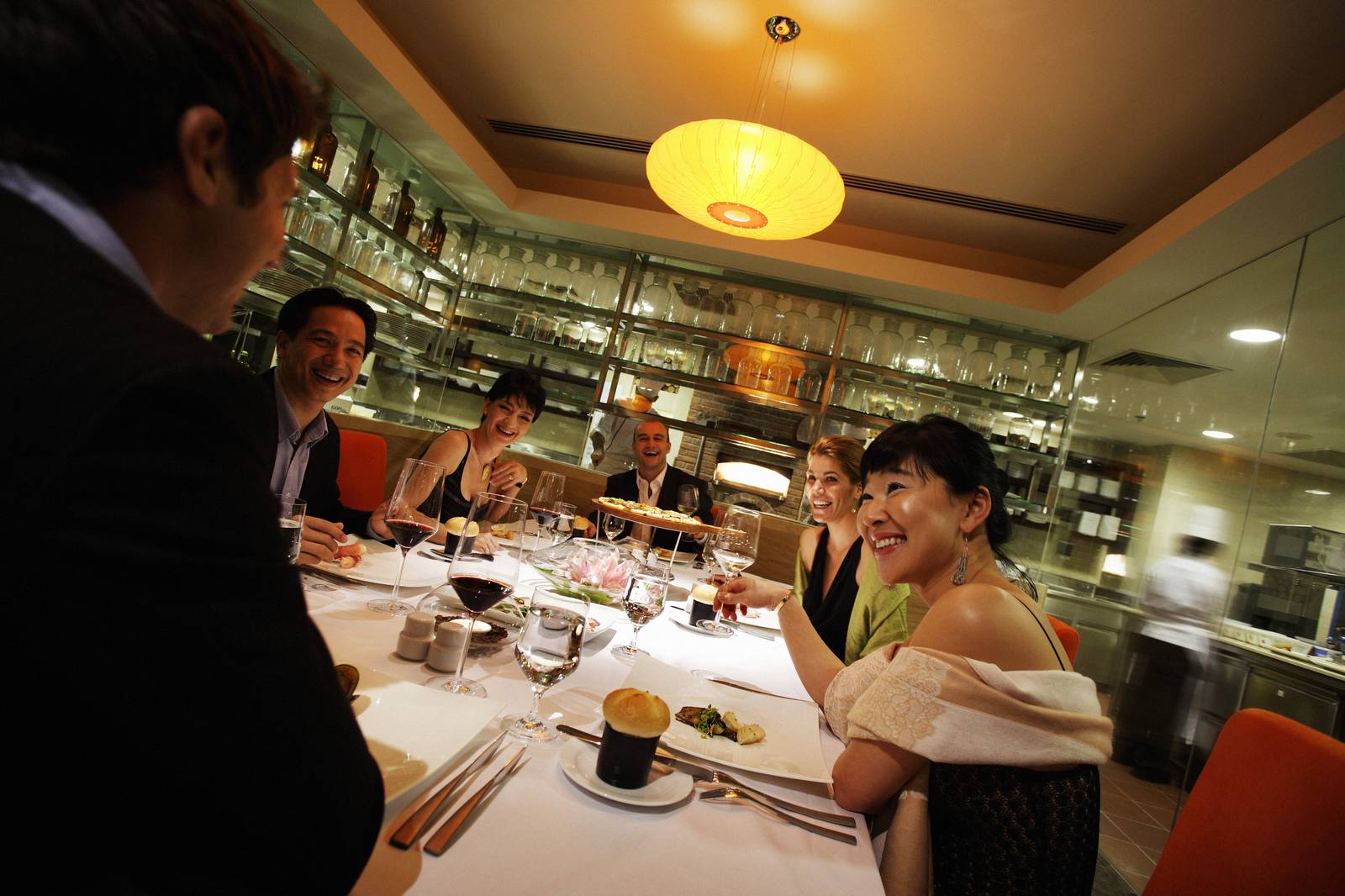 河內西湖洲際酒店 Milan 餐廳長桌，白桌布、紅酒杯與中央薄餅，後方玻璃酒架與磚拱柴燒窯
