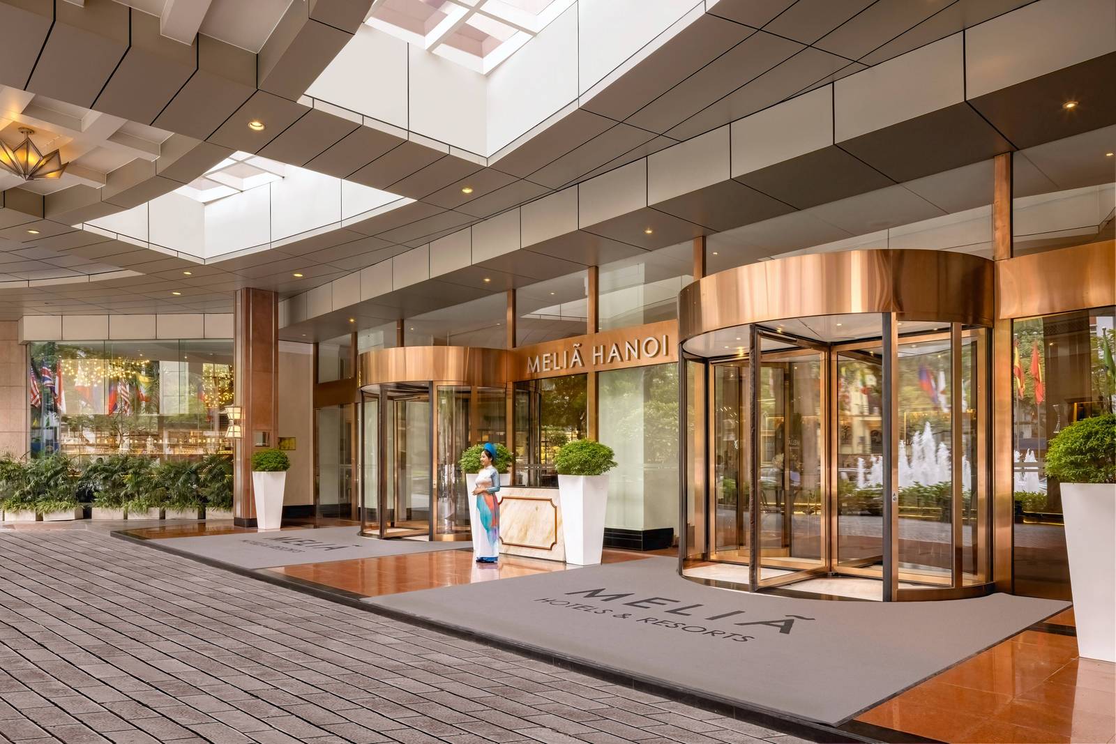 河內美利亞酒店入口:兩座銅色迴轉門、上方 MELIÁ HANOI 金字,中間講台站著穿白襖戴頭巾的接待人員
