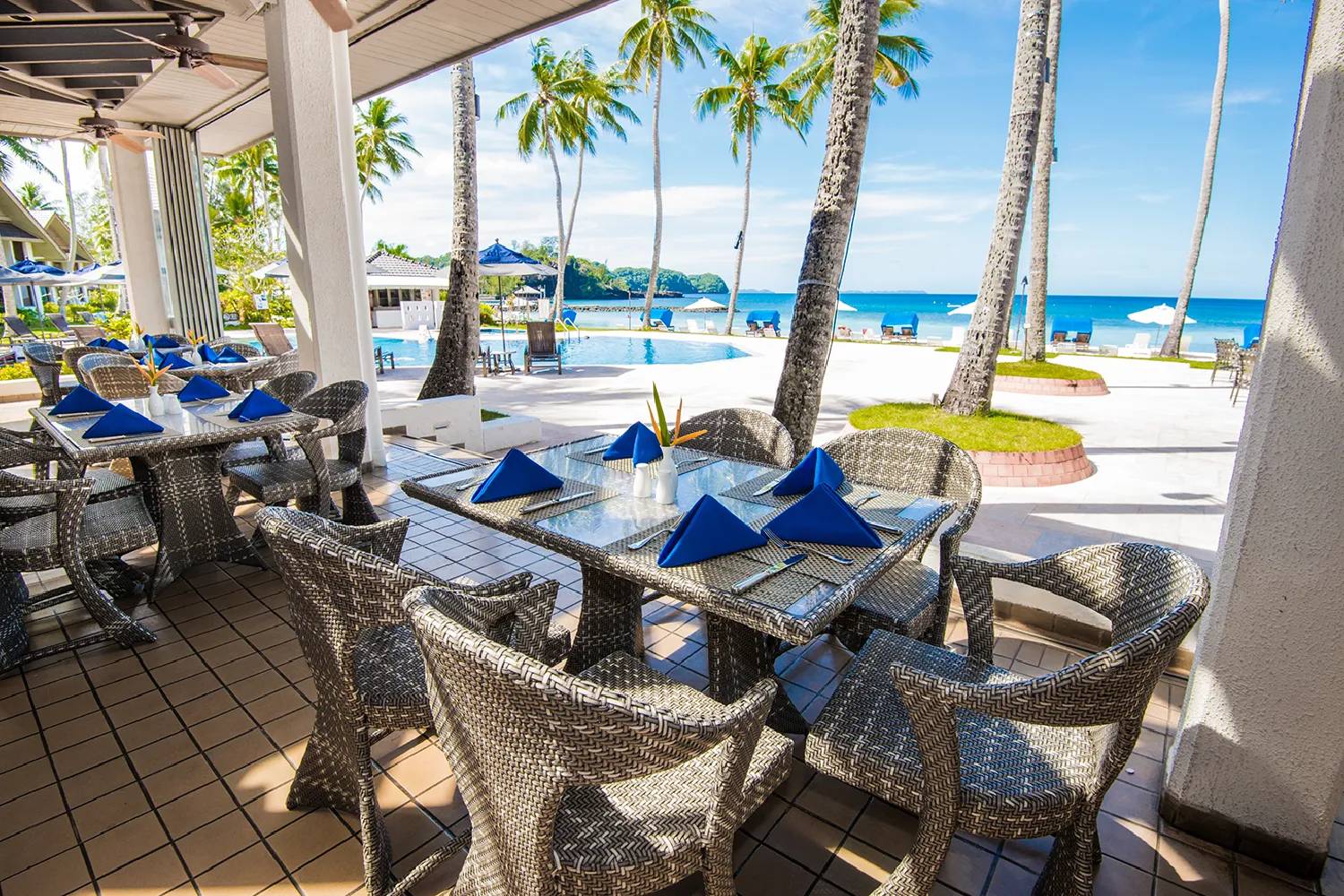 Coconut Terrace 露台餐桌、藍色餐巾、泳池、棕櫚與後方白沙海灘