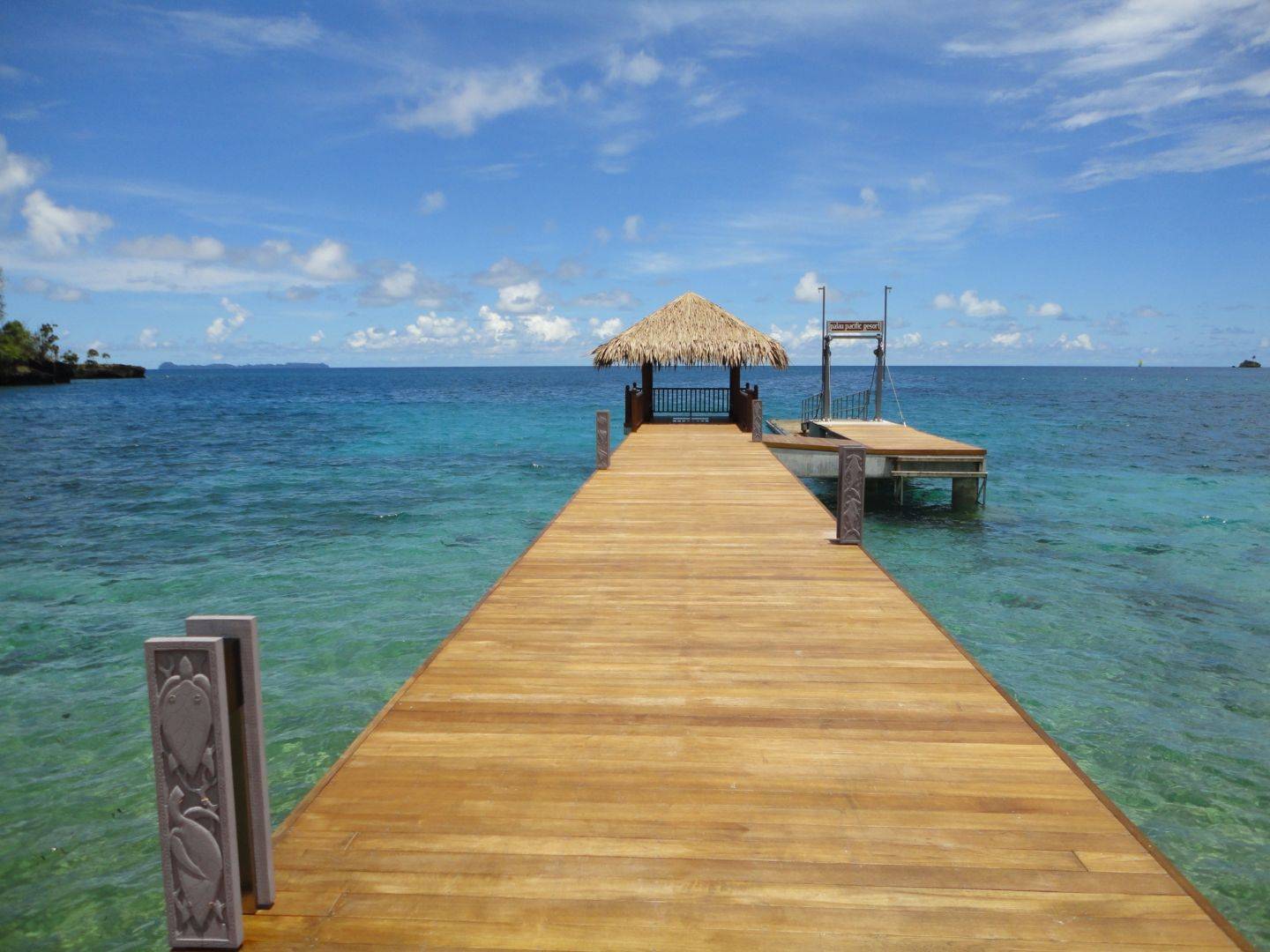 木棧橋伸入清澈淺灣,盡頭茅草亭與 palau pacific resort 字樣招牌
