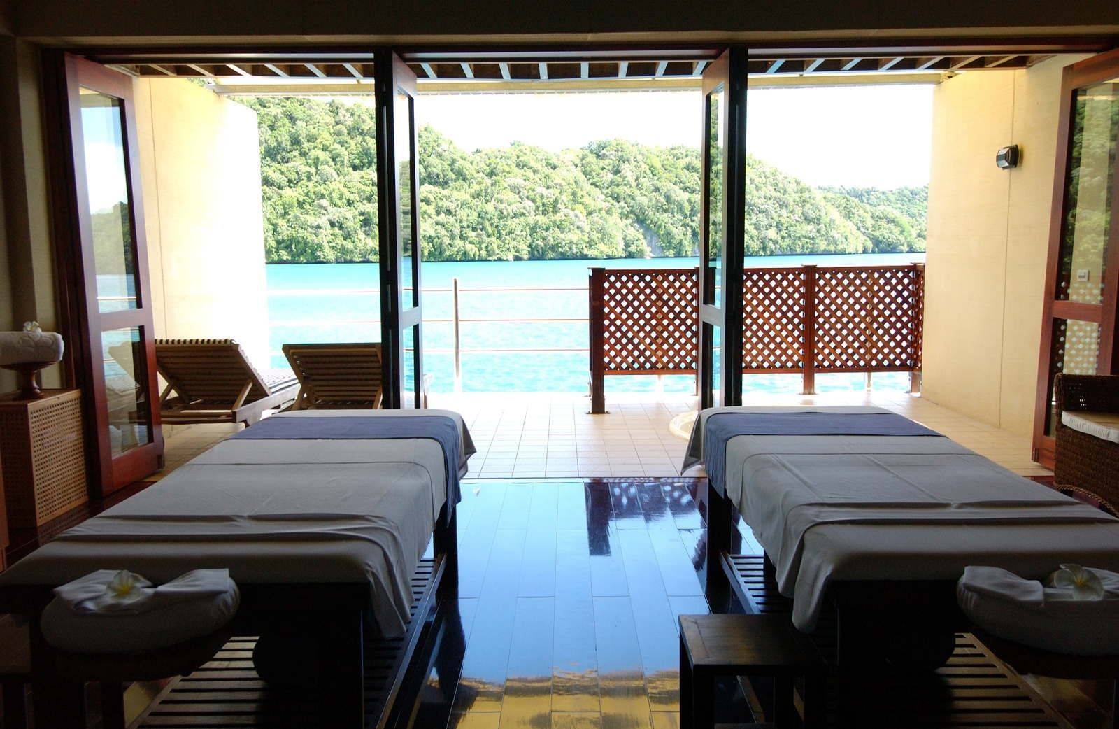帛琉老爺大酒店 Mandara Spa 雙人治療室:兩張按摩床面向陽台,窗外是碧水與林木覆蓋的島丘