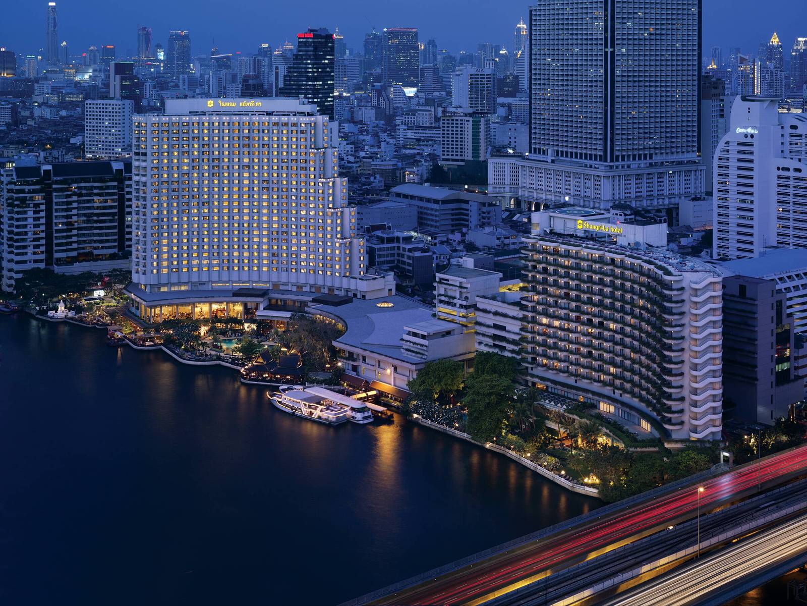 曼谷香格里拉酒店夜景：Shangri-La Wing 與曲線的 Krungthep Wing 亮燈臨昭披耶河，碼頭停白色河船，前景高架橋車軌光跡