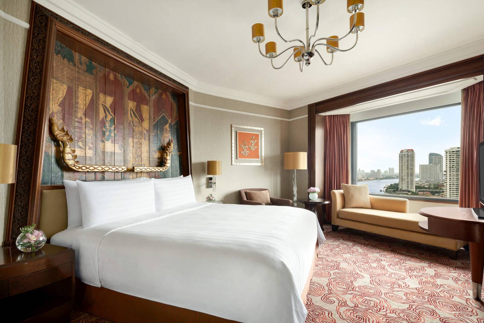 曼谷香格里拉酒店 Executive River View Suite：大床上方深色木框泰式彩繪壁飾與金色浮雕，窗邊貴妃椅，窗外是昭披耶河與對岸建築