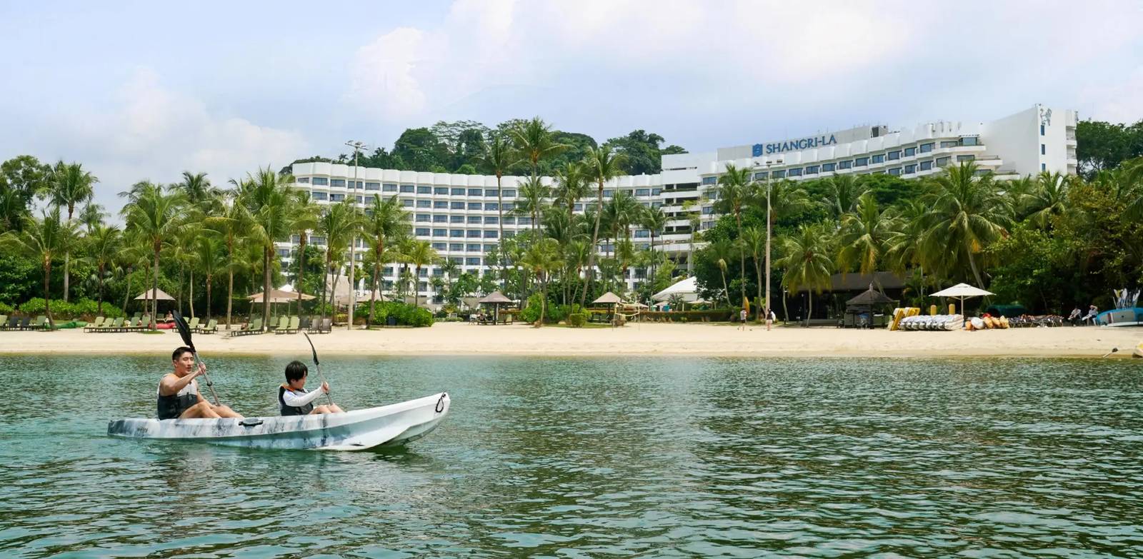 從海上望向新加坡聖淘沙香格里拉度假村：前景雙人獨木舟，後方是西羅索沙灘、棕櫚與標有 Shangri-La 的弧形白樓