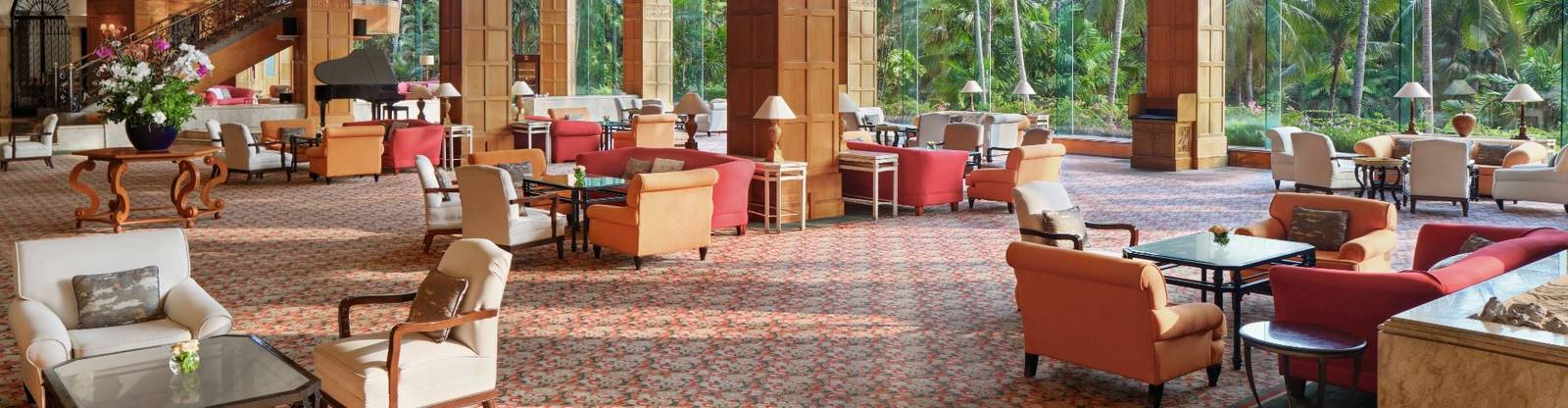 泗水香格里拉酒店 Lobby Lounge 有奶油與紅橘色扶手椅、木柱與樓梯，落地窗望向棕櫚庭園，後方可見黑色平台鋼琴