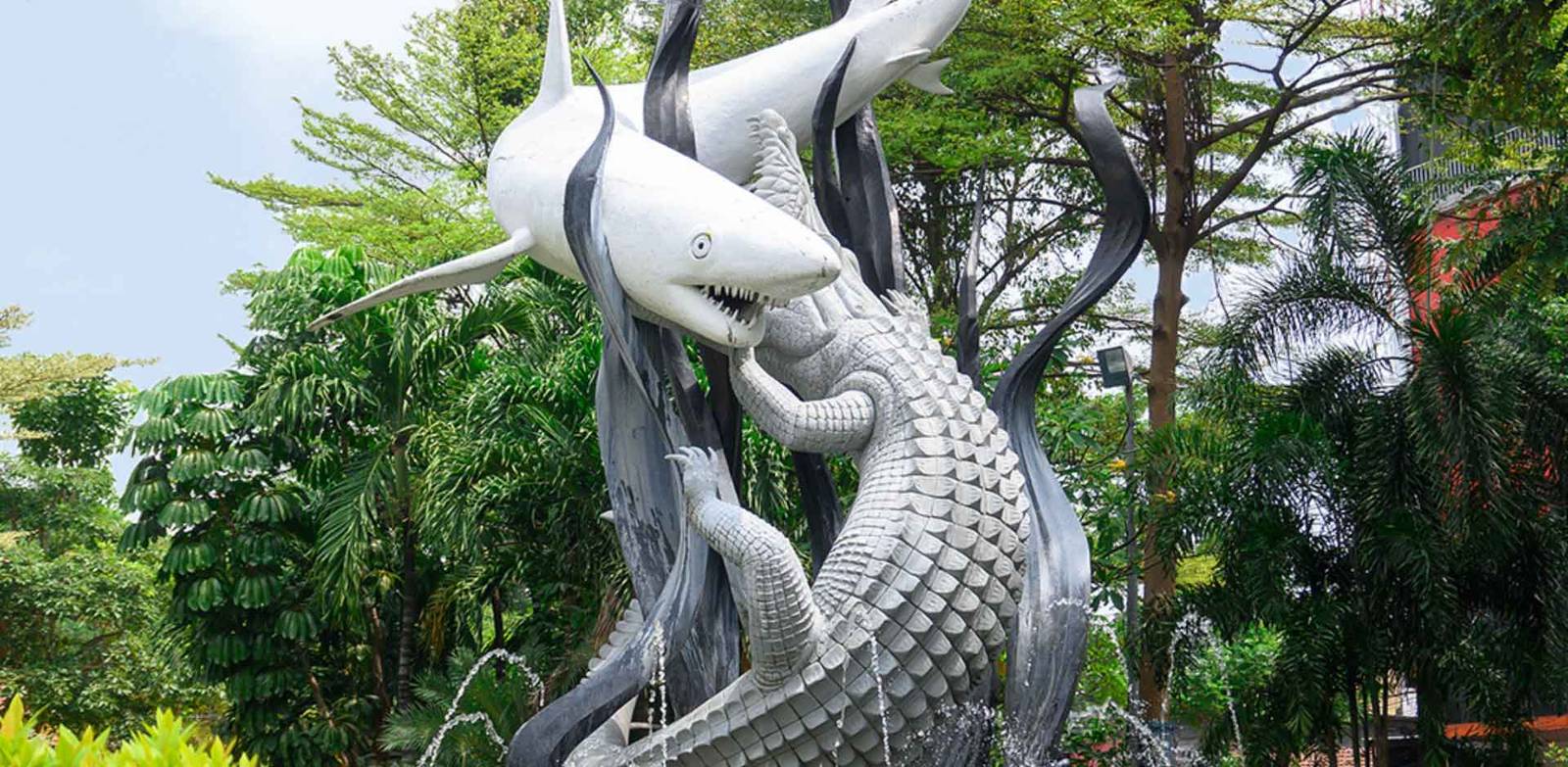 泗水地標蘇拉與巴亞雕像：白色鯊魚與灰色鱷魚交纏在噴泉座上，後方是熱帶樹叢。此圖為城市象徵，不是飯店園區