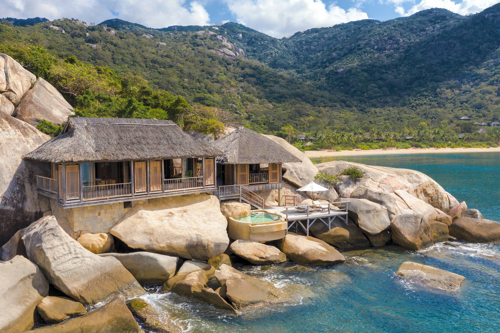 花崗岩岸上的木造茅草頂別墅，私人泳池與木甲板緊貼碧藍海水