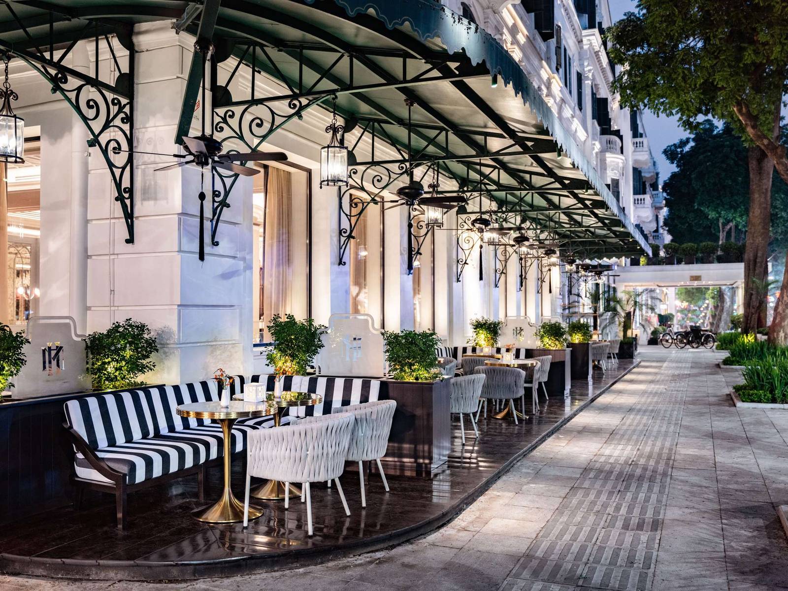 河內索菲特傳奇大都會酒店臨街露台 La Terrasse,綠雨篷下黑白直條沙發與大理石圓桌,外側是人行道與街樹