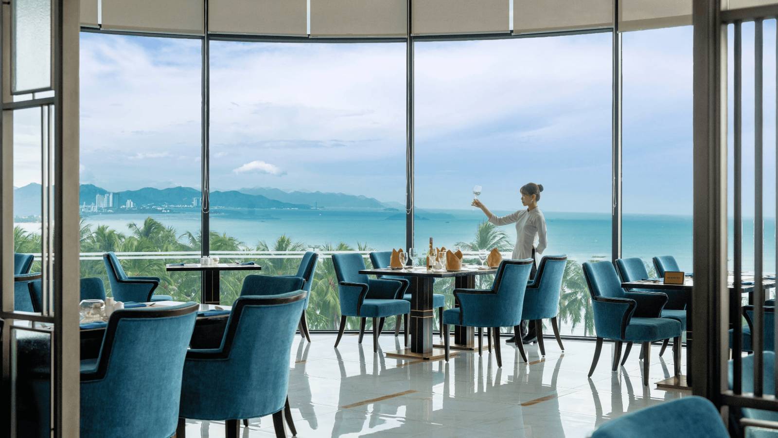 芽莊珍珠海濱酒店 Coral 餐廳藍綠色座椅與落地窗外的棕櫚樹梢、海灣與遠山