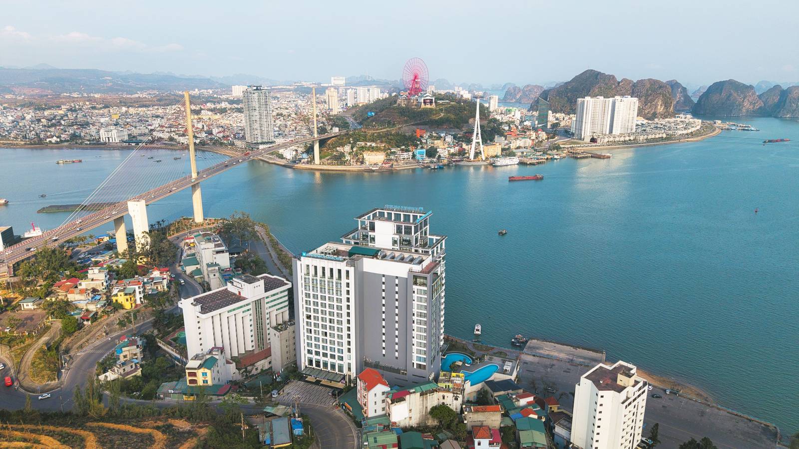 下龍灣溫德姆傳奇酒店日間空拍,白色雙塔臨水,左後方白齋大橋、山丘太陽之輪與遠方下龍灣石灰岩島