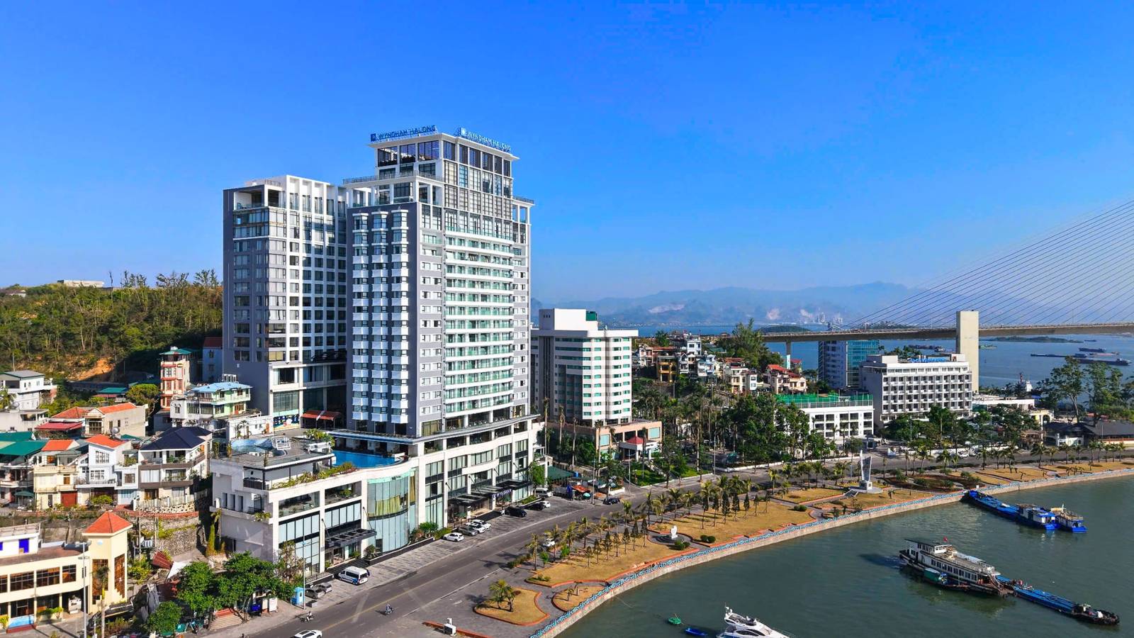 下龍灣溫德姆傳奇酒店日間高角度外觀,樓頂 WYNDHAM LEGEND 字樣、層層陽台,右下沿街泊船與白齋大橋