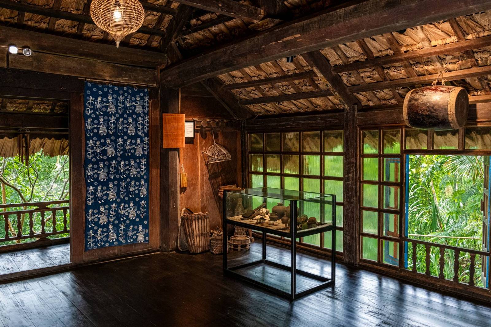 木造高腳屋內的藍色織布、展示櫃與木窗外的綠意
