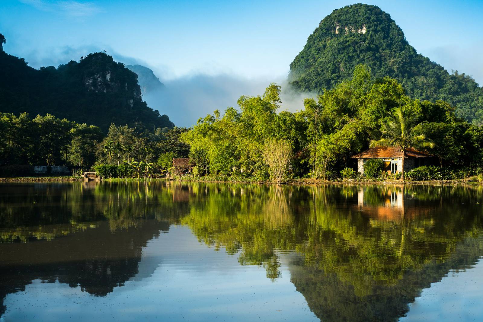 晨霧中的石灰岩山與湖面倒影，岸邊有一棟紅瓦小屋