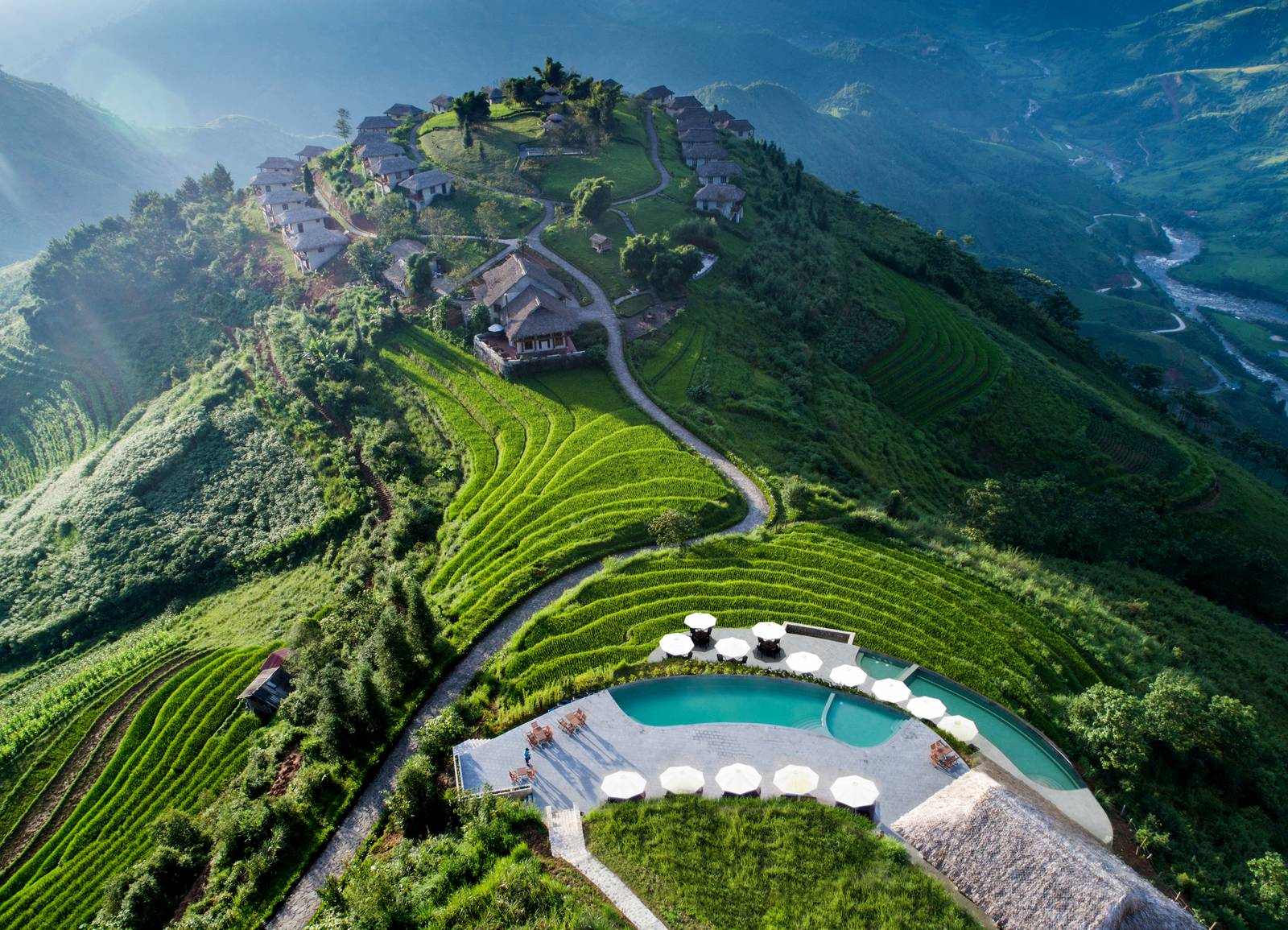 空拍山頭上的茅草小屋群、彎曲山路、梯田與白色陽傘圍繞的泳池